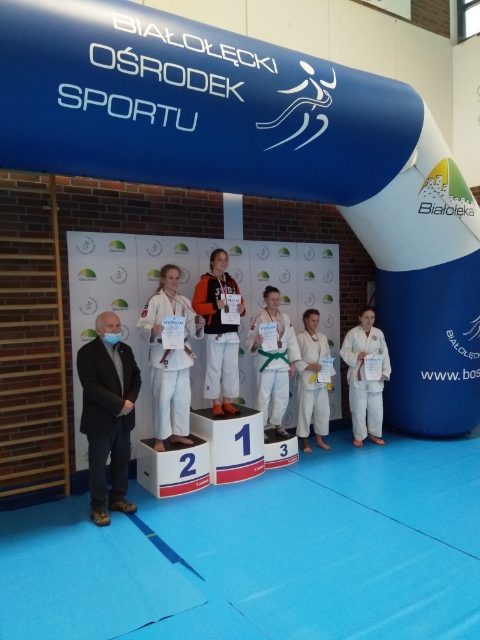 grupa nagrodzonych judoków - trzech stoi na podium, dwóch obok podium, wraz z nimi organizator