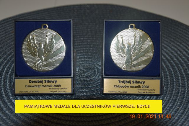 Na zdjęciu na szarej podkładce leżą dwa granatowe pudełka z medalami w kolorze srebrnym. Pod spodem na żótym tle napis: Pamiątkowe medale dla uczestników pierwszej edycji.