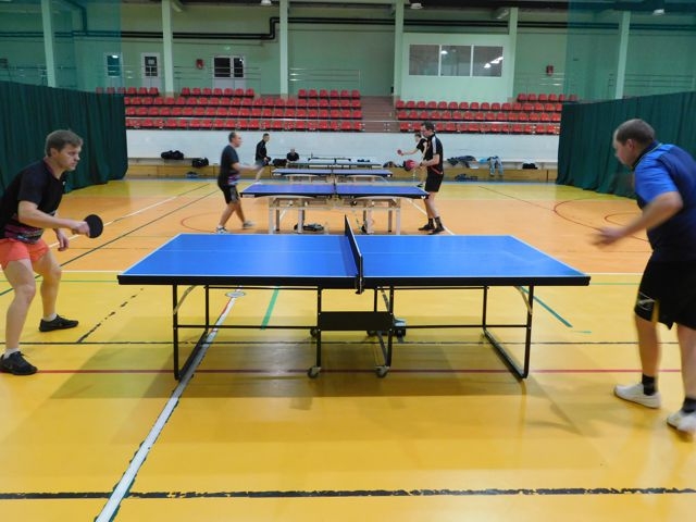 W centrum zdjęcia stoi stół do tenisa. Z obu stron znajdują się zawodnicy podczas gry.