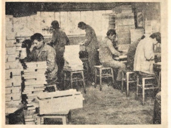  skan czarno-białego zdjęcia prasowego, na którym widoczna jest pracownia stolarska; widocznych jest sześciu pracowników przy stołach oraz stosy pudłem i innych elementów drewnianych