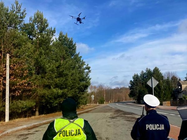 dwóch policjantów stoi przy drodze i sterują dronem, ktory wzniósł się w powietrze
