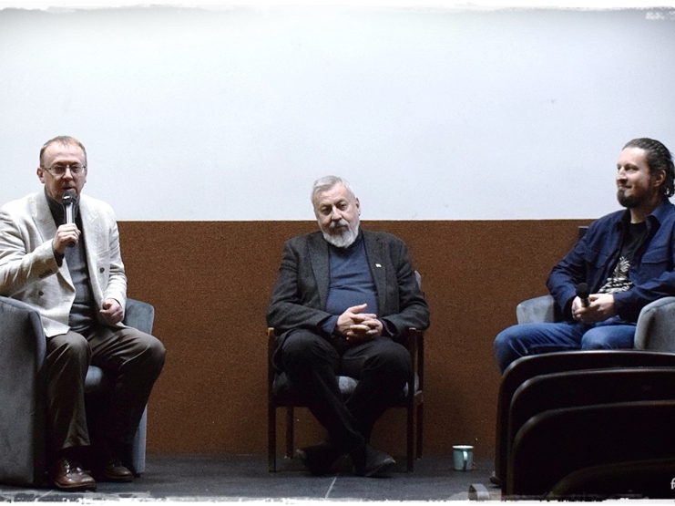 Trzej mężczyźni siedzą na fotelach. Mężczyzna po lewej przemawia do mikrofonu.