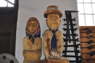 dwie drewniane rzeźby, mężczyzny i kobiety, pod ścianą stoi oparte dawne narzędzie gospodarskie służące do uprawy ziemi