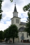 budynek kościoła w kolorze białym ze strzelistą wieżą w otoczeniu zielonych drzew