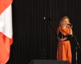kobieta w pomarańczowej sukience, wypowiadająca się do mikrofonu; z lewej strony widnieje fragment biało-czerwonej flagi