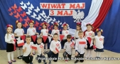 Dzieci ubrane na galowo trzymają biało-czerwone flagi, za nimi napis wiwat maj 3 maj.
