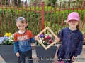 Dzieci kucają i przez drwenianą ramkę pokazują kwiaty w donicy.