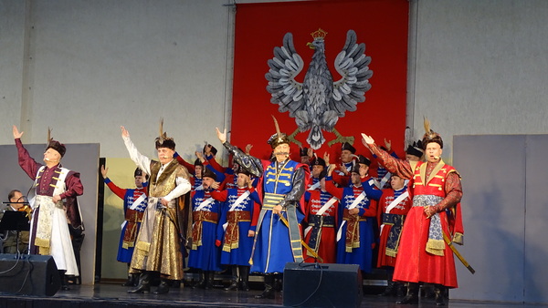 na scenie amfiteatru w Hajnówce stoi grupa artystów w strojach historycznych. w tle widać duże godło Polski