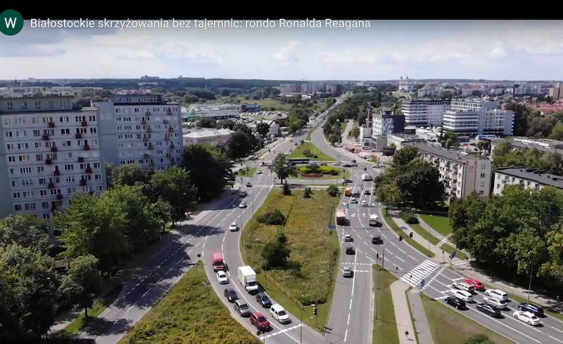 w środkowej części przebiegają ulice z licznymi samochodami. Z lewej strony wysokie bloki. Na zdjęciu widać dużo zieleni miejskiej.