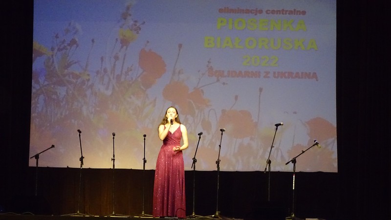 laureatka w czerwonej sukni śpiewa na scenie. W tle wyświetlony został duży slajd z tutułem konkursu