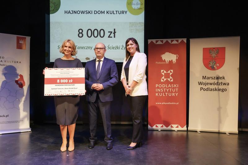 dwie kobieta i mężczyzna pozują do zdjecia na scenie. Kobieta po lewej trzyma czek z dotacją na 8 tys zł
