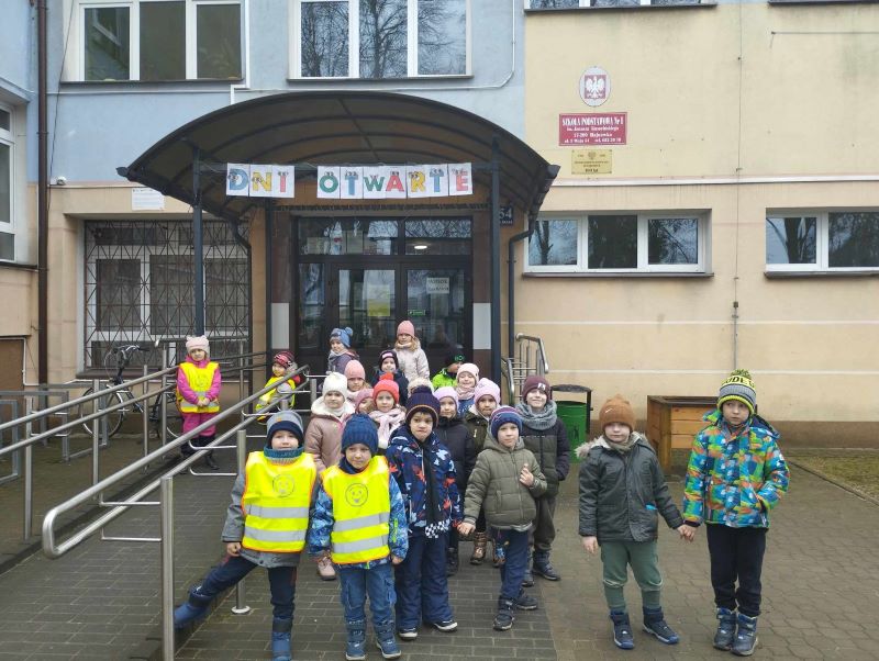 Dzieci przed budynkiem szkoły. Nad drzwiami wisi napis DNI OTWARTE