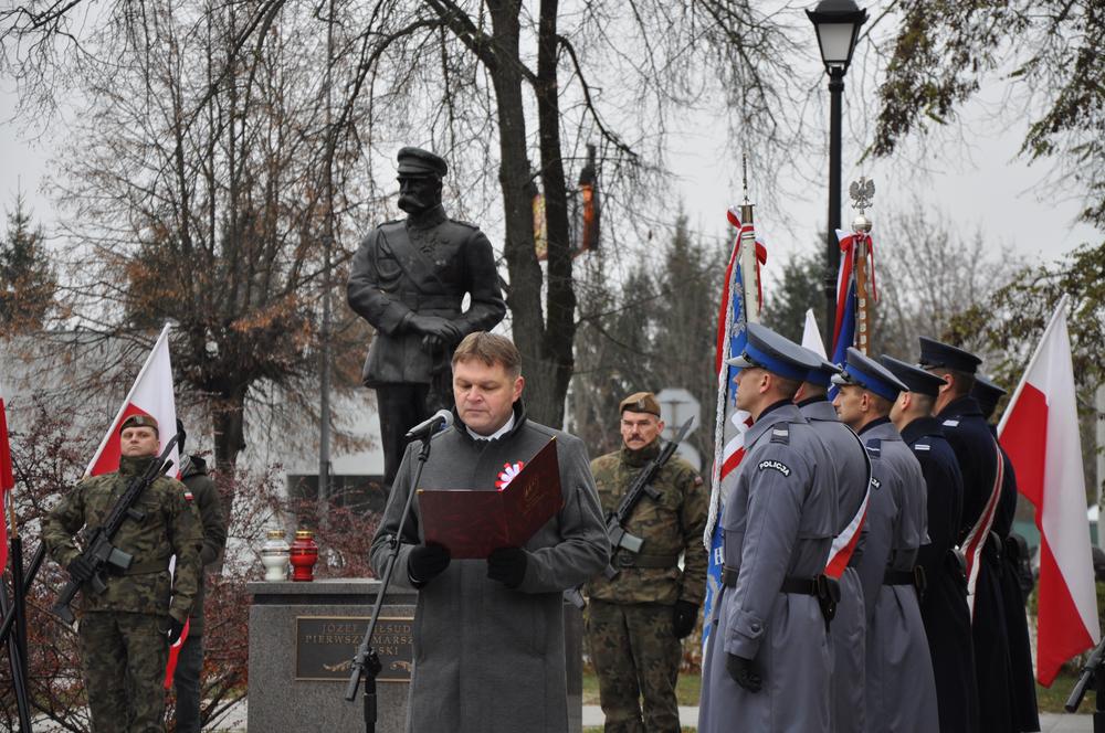 Mężczyzna stoi przy mikrofonie. Obok stoją poczty sztandarowe, w tle widać pominik Marszałka JózefaPiłsudskiego