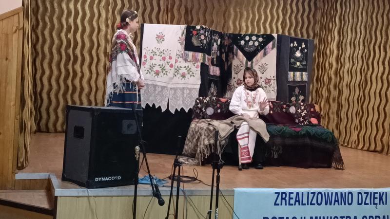 dwie osoby w stojach ludowych na scenie. W tle widać zawieszone haftowane makaty i chusty