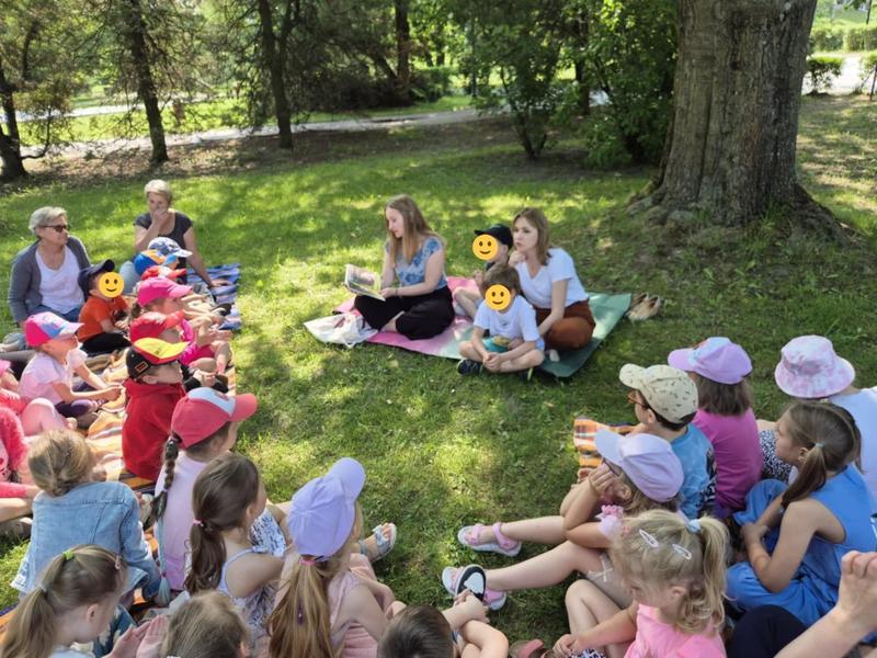 dzieci siedzą na polanie i słuchaja opowieści czytanej przez opiekuna