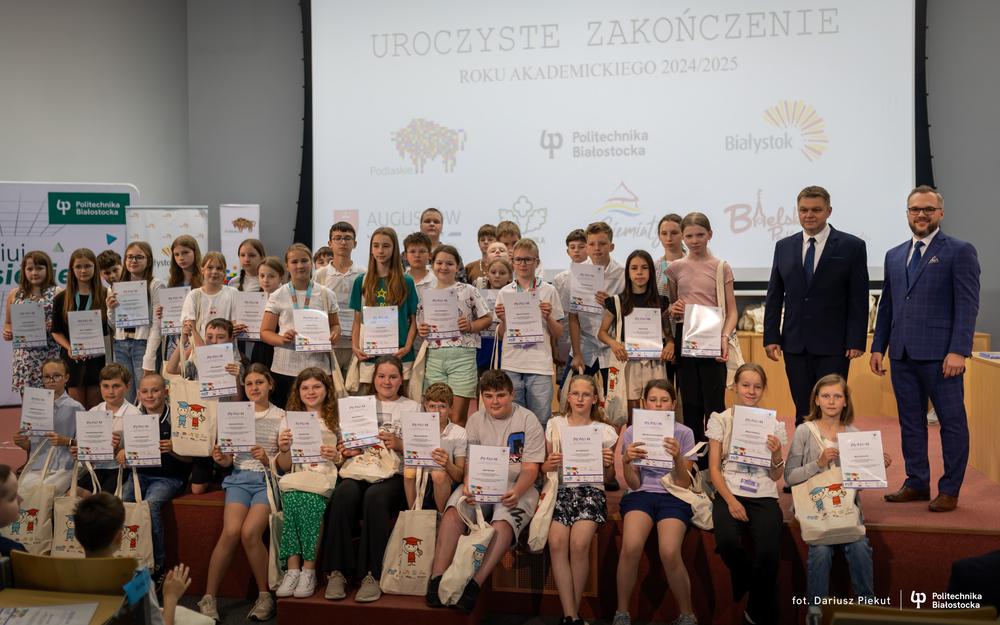 duża grupa dzieci pozuje do wspólnego zdjecia z Burmistrzem Miasta Hajnówka i Prorektorem ds. Studenckich Politechniki Białostockiej. Dzieci trzymają dyplomy zakończenia roku akademickiego.