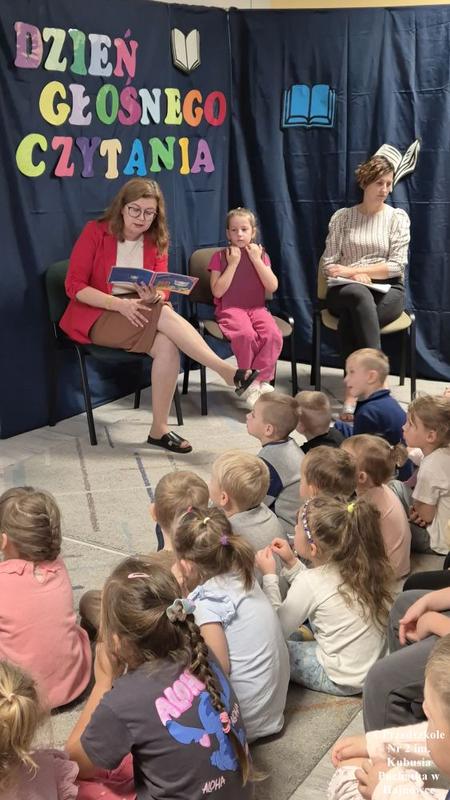 trzy osoby siedzą przy ściance z napisem DZIEŃ GŁOŚNEGO CZYTANIA. Pani Dyrektor czyta z książki.