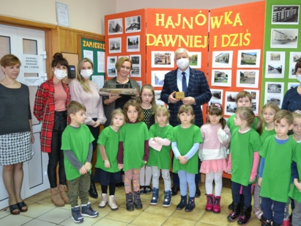 Dzieci, kadra pedagogiczna oraz delegacja z URzedu Miasta pozują do grupowego zdjecia. Z tyłu widać gazetkę ścienną ze zdjeciami Hajnówki dawniej i dziś