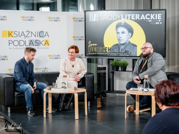 trzy osoby siedzą przy stolikach. W tle wyświetlane jest zdjecie autora pamiątnika