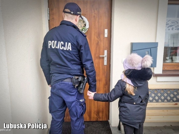 Umundurowany policjant trzyma za rękę dziecko. Oboje stoją przed drzwiami domu.
