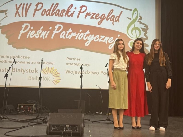 trzy młode kobiety stoją na scenie. W tle wyświetlany tytuł konkursu
