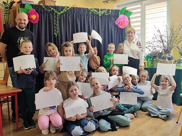grupa dzieci pokazuje swoje prace. Obok stoi opiekunka i prowadzący zajęcia