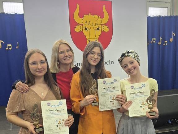 dziewczęta ze statuetkami i dyplomami pozują z instruktorką do zdjęcia. W tle herb Tykocina