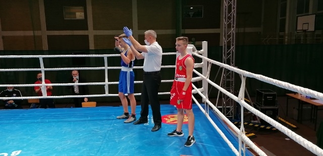 W ringu stoi dwóch zawodników, jeden z nich ubrany w czerwony, a drugi w niebieski strój bokserski. Pomiędzy nimi stoi sędzia z maseczką na twarzy.