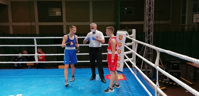 W ringu stoi dwóch zawodników, jeden z nich ubrany w czerwony, a drugi w niebieski strój bokserski. Pomiędzy nimi stoi sędzia z maseczką na twarzy.