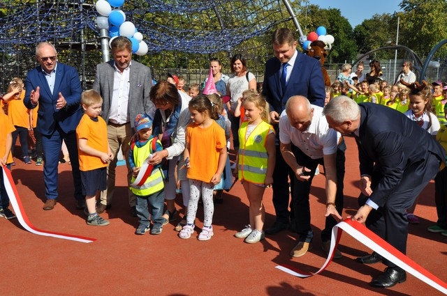 władze miasta, powiatu, domu kultury otwierają plac zabaw poprzez symboliczne przecięcie biało-czerwonej wstęgi; wokól nich stoją dzieci