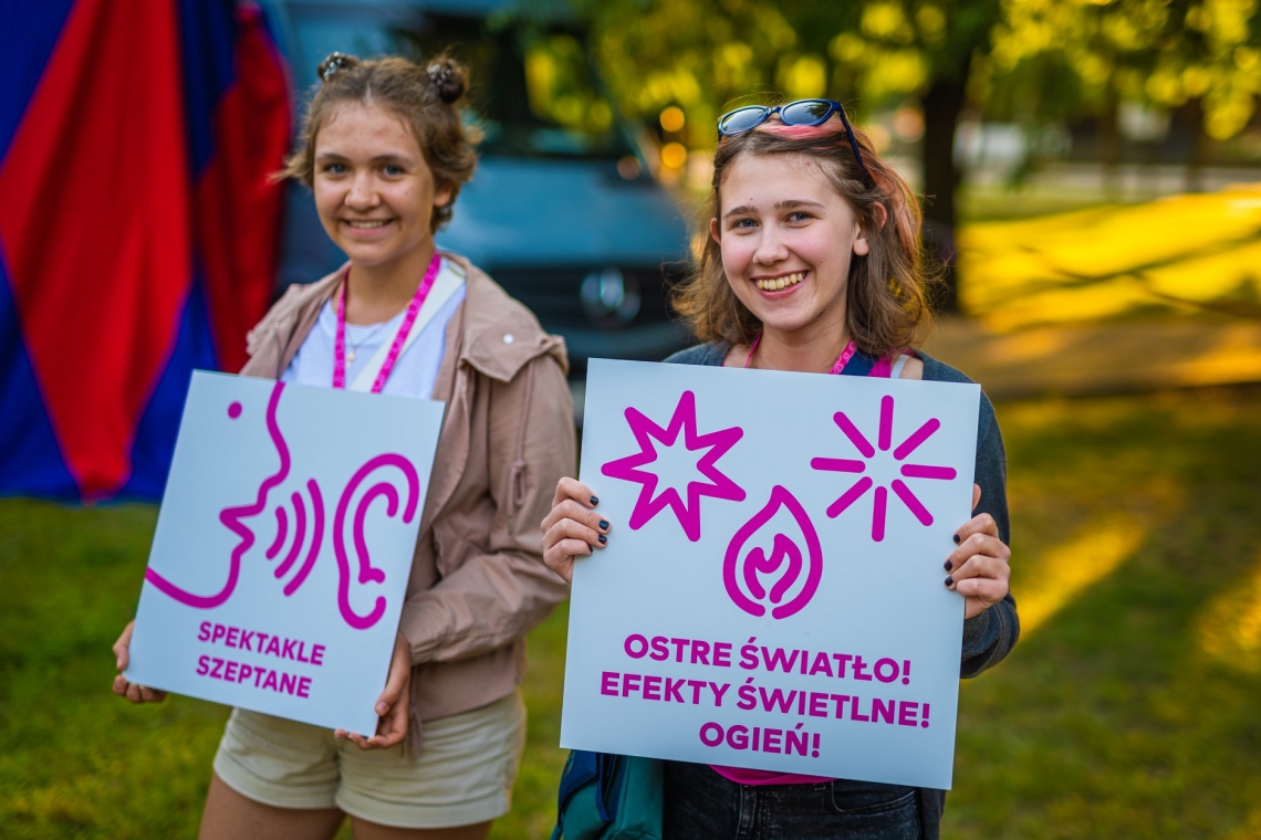 Na zdjęciu stoją dwie młode dziewczyny. Jedna z nich trzyma tabliczkę z napisem: Spektakle szeptane, druga: ostre światło! efekty świetlne! ogień!. Każdy rysunek ma obrazujący napisy piktogram.