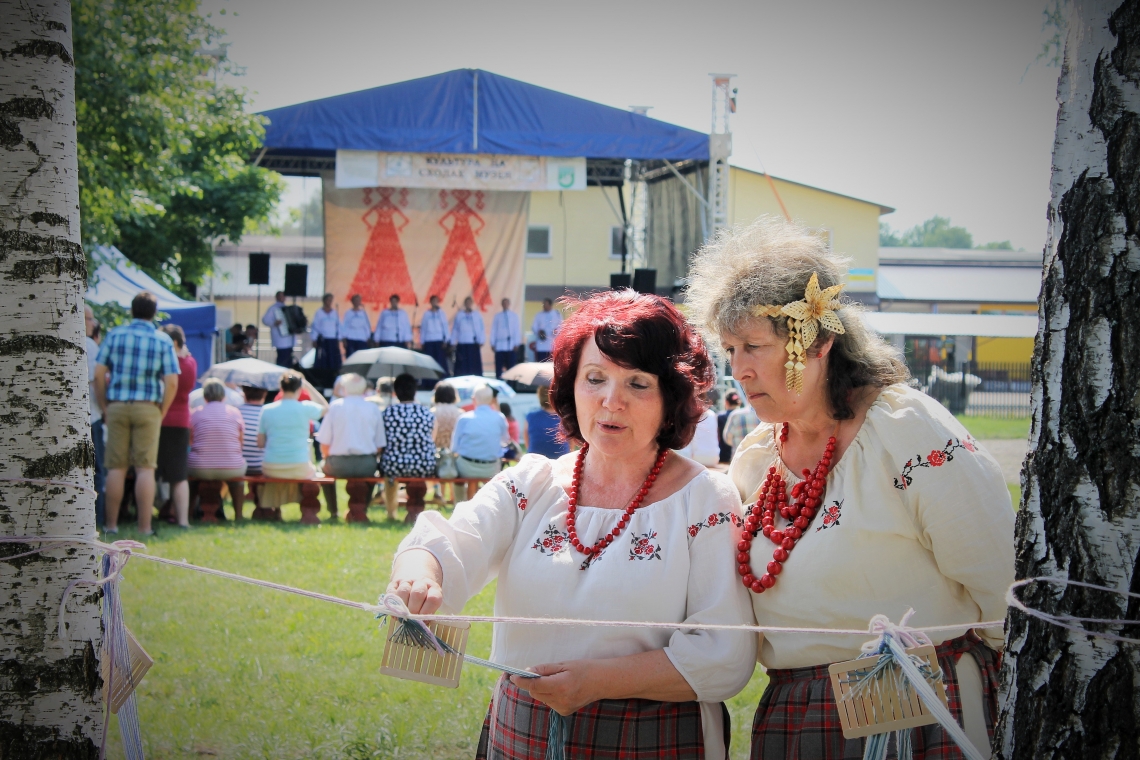na pierwszym planie dwie kobiety w strojach ludowych, na szyi mają czerwone korale; w tle scena z niebieskim zadaszeniem, przed sceną na ławkach siedzą ludzie, wysłuchujący występujących artystów