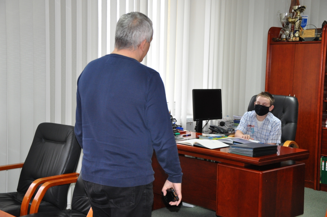 za biurkiem siedzi kilkunastoletni chłopiec; do gabinetu wszedł mężczyzna w granatowej bluzce, stoi przodem do chłopca; z lewej strony petenta stoją dwa czarne skórzane fotele