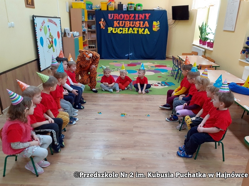 Zdjęcie ukazuje dzieci siedzące na krzesełkach, które obserwują i dopingują, jak dzieci biorą udział w zabawie sprawnościowej „Wyścig pszczółek”.