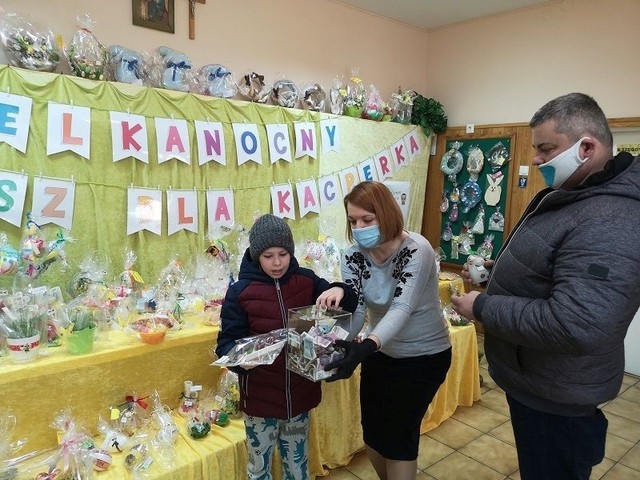 Na zdjęciu widać ojca z synem i jedną z organizatorek na tle wystawy. Chłopiec wrzuca pieniądze do skrzynki, którą trzyma nauczycielka.