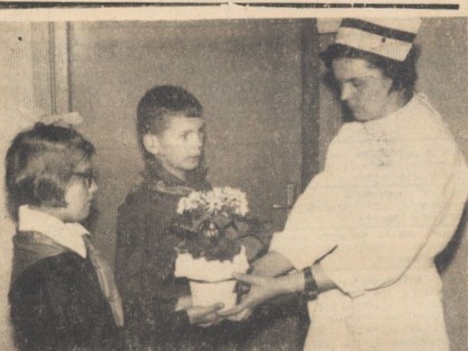 skan czarno-białego zdjęcia prasowego; na zdjęciu stoją po lewej chłopiec i dziewczynka, którzy wręczają kwiaty szkolnej higienistce, stojącej po prawe