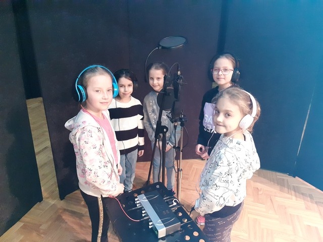 Pięć dziewcząt stoi przy sprzęcie nagrywającym. Na głowach mają założone słuchawki.