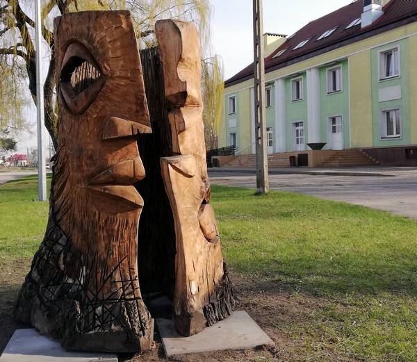 Na tle budynku HDK stoi rzeźba w drewnie, która przedstawia dwie strony ludzkiej głowy. Jedna z części stoi do góry nogami.