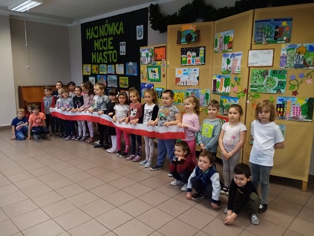 Dzieci trzymają biało-czerwoną wstęgę na tle tablic z pracami.
