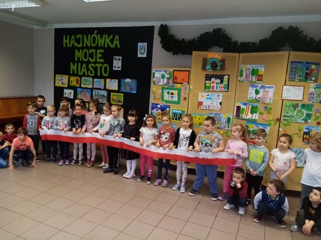 Dzieci trzymają biało-czerwoną wstęgę na tle tablic z pracami.