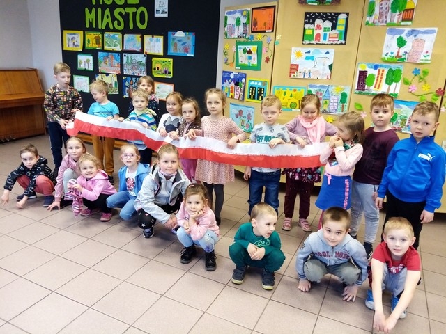 Dzieci trzymają biało-czerwoną wstęgę na tle tablic z pracami.