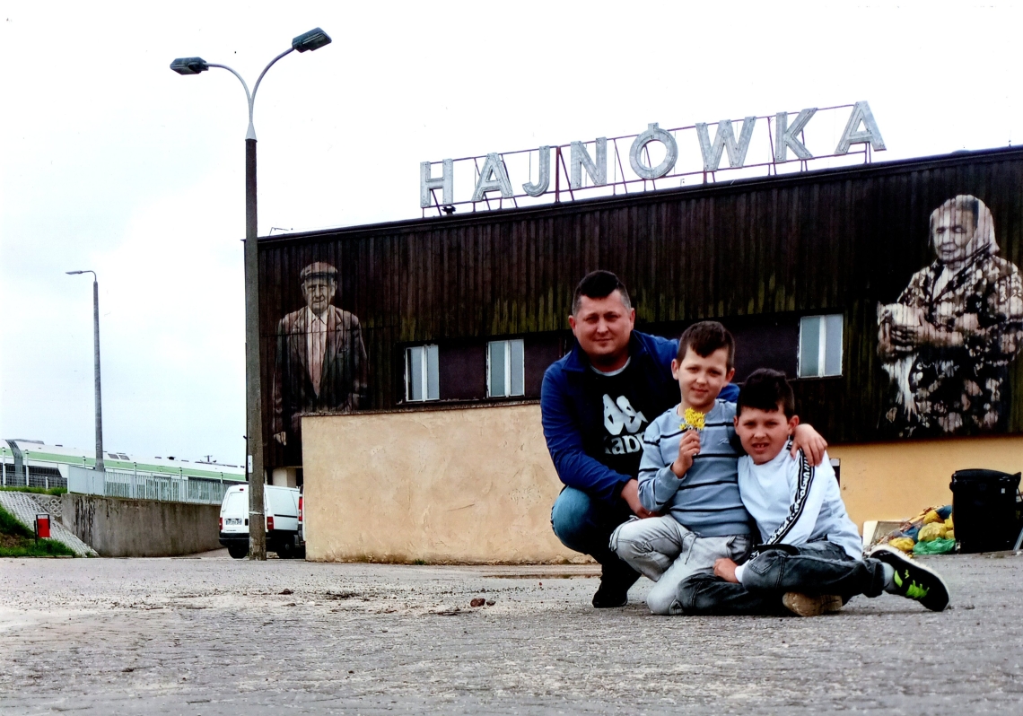 dwóch chłopców wraz z ojcem pozują przykucnięci na tle budynku dawnego dworca; widoczna jest ściana z muralem starszego mężczyzny i kobiety oraz duży napis Hajnówka na dachu