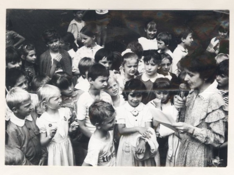 grupa dzieci stoi wokół kobiety z mikrofonem i plikiem kartek