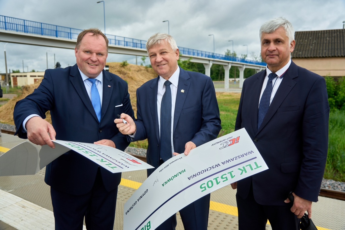 Trzech mężczyzn stoi obok siebie, dwóch z nich trzyma  w rękach pamiątkowy bilet w kształcie prostokąta.