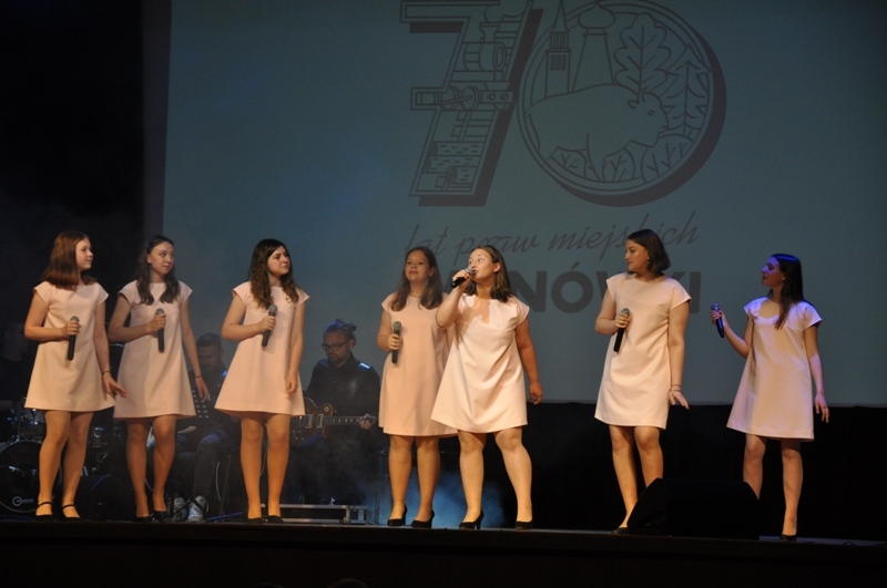 7 dziewczyn w białych sukienkach na scenie wykonują piosenkę