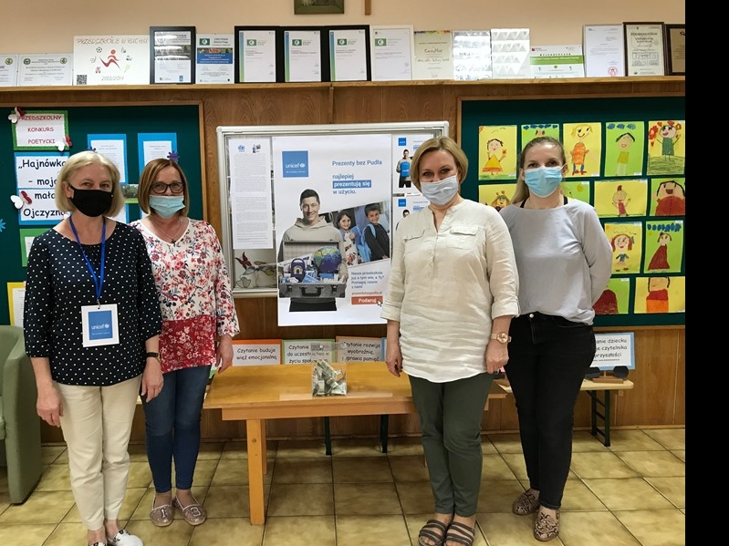 Zdjęcie przedstawia Panią Dyrektor wraz z nauczycielkami – wolontariuszkami UNICEF w trakcie kwesty na zakup artykułów humanitarnych. Wolontariuszki stoją na tle plakatów UNICEF „Prezent bez Pudła”