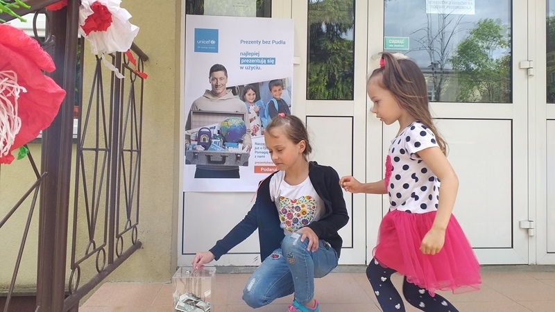 dzieci na tle plakatu UNICEFU, każde z nich wrzuca do puszki pieniądze.