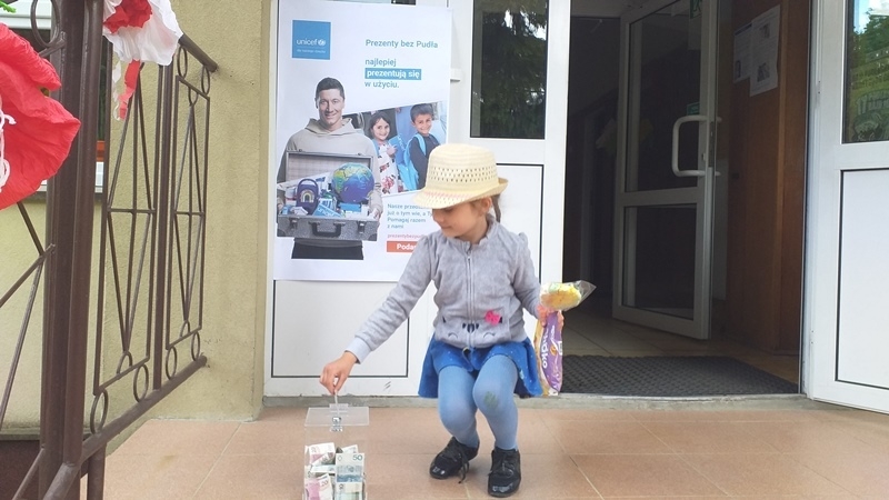 dzieci na tle plakatu UNICEFU, każde z nich wrzuca do puszki pieniądze.