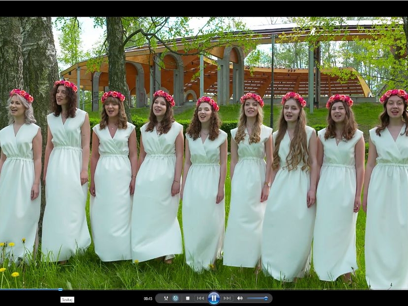 9 dziewczyn w białych długich sukienkach z różowanymi wiankami na głowie, w otoczeniu zieleni traw i drzew