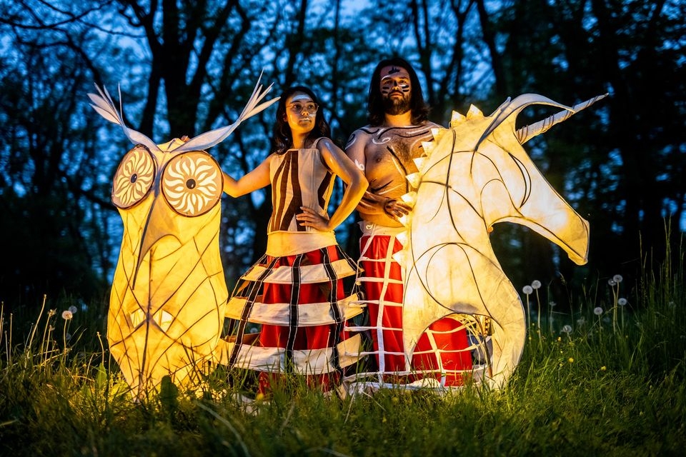 dwaj aktorzy, kobieta i mężczyzna; obok nich stoją dwa podświetlone lampiony w formie zwierząt; stoją na zielonej trawie pod osłoną nocy